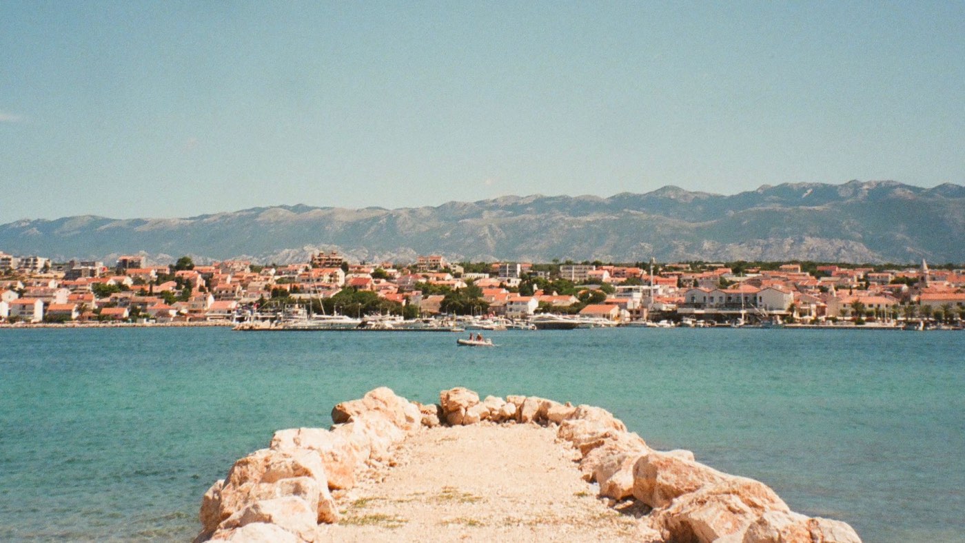 A Croatian coastal town seen across turquoise water from the end of a rocky breakwater, with a mountain range rising behind the rooftops